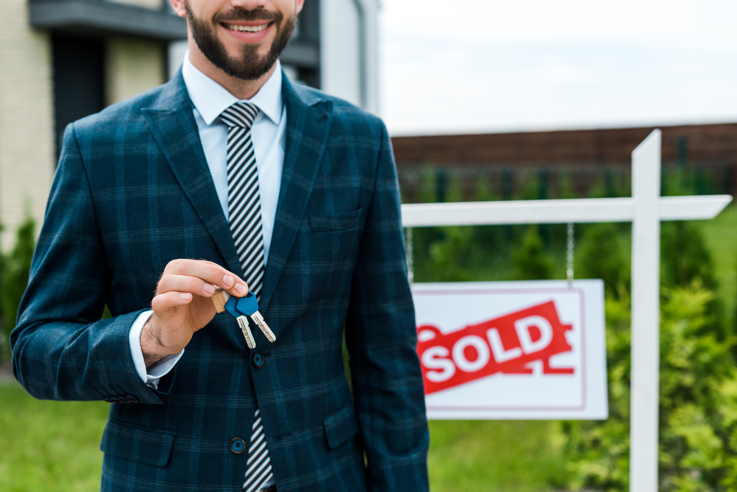 Young male realtor holding house keys in front of a sold sign